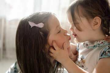 Two cute sisters of preschooler age pull their lips together to kiss each other in dreamy soft backlight. The girl holds her sister's rosy cheeks with her small hands and looks at her dreamy face.