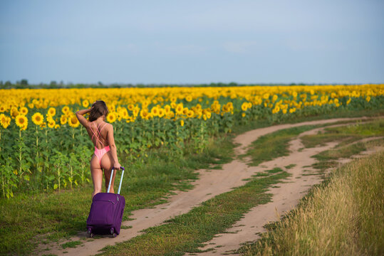 Beautiful woman in a pink swimsuit travels through the countryside with a suitcase. A girl in a monokini is walking alone on a field of sunflowers with a big bag. Hiking. Rest and social distance.