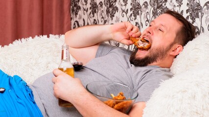 Obraz na płótnie Canvas fat man drinks beer and eats unhealthy food chicken wings, bored in front of the TV outlook on the couch. The concept of malnutrition, quarantine at home, alcoholism.