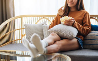 Closeup image of a young woman holding and eating pop corn while watching tv and sitting on sofa at home