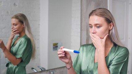 Shocked woman holding positive pregnancy test in bathroom