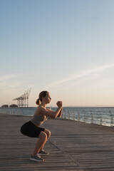 the girl is engaged in fitness by the sea or ocean, crouches and smiles, in the morning light the concept of an active lifestyle.