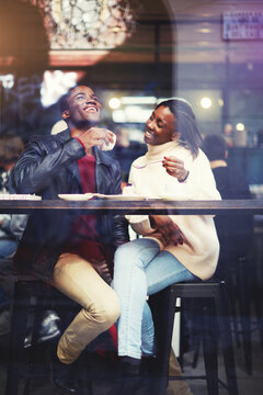 View through window of young beautiful couple in love drinking coffee and laughing while sitting in coffee shop interior, happy black man and woman having a great time together while relaxing in cafe
