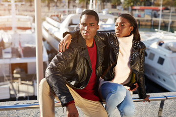 Portrait of fashionable professional couple of models posing outdoors, embracing black couple enjoying time spending together while sitting in yacht port of Barcelona, vacation holidays
