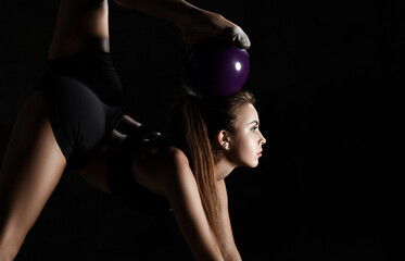 Young sporty woman doing gymnastics stretching fitness exercises workout with purple ball in gym sport club on dark 