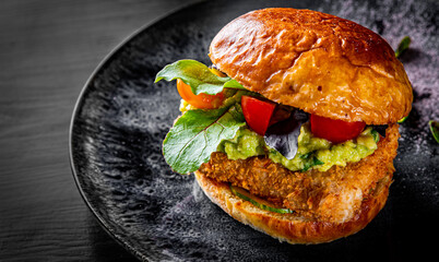fish burger with cheese, guacamole sauce and vegetables in plate on black wooden table background