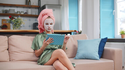 Woman with facial beauty mask using tablet in living room