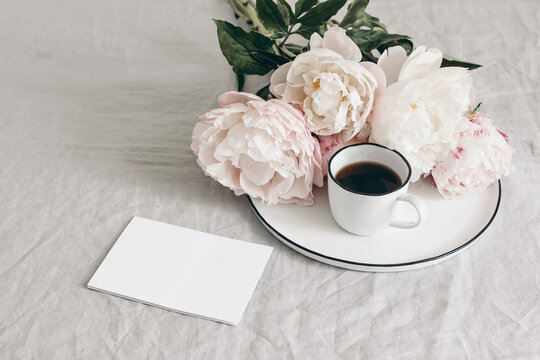 Spring breakfast still life scene. Cup of coffee and bouquet of white and pink peonies on porcelain plate. Blank greeting card mockup on linen table cloth background. Vintage feminine photo. No people