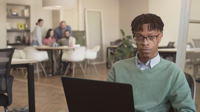 Lockdown portrait of young African man sitting in coworking space, working on PC then looking at camera while group of four people communicating on background