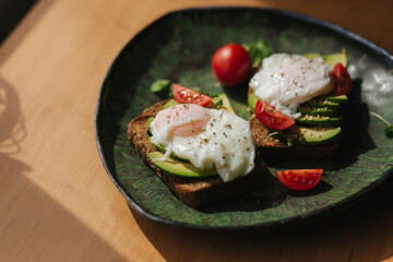Delicious breakfast at home. Sandwich with fresh sliced avocado above rye toasted bread with cherry tomatoes and poached egg on green plate. Top view