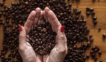 Hands of an old woman holding coffee beans over the table on which the coffee beans are lying.