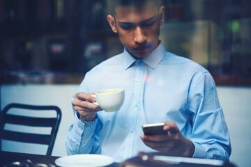 Attractive concentrated young student dressed in trendy shirt spending leisure time on break in cafeteria monitoring latest news on informational websites using free internet access in coffee shop