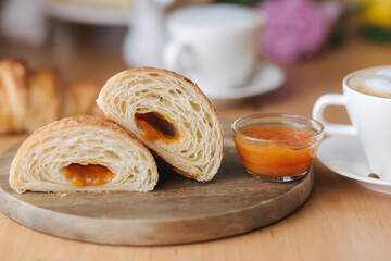 Two halves of freshly bared croissant on wooden board with delotious homemade apricot jam. Background of cappuccino. Morning breakfast