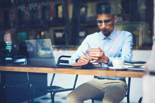 Handsome young male international student in trendy outfit and eyewear preparing to exams in university solving problems and making practising tasks sitting in cafe using free internet connection