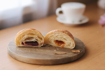 Two halves of freshly bared croissant on wooden board with different jam. Background of cappuccino