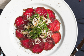 Flat lay of beef carpaccio in on the white plate with arugula, mushrooms, Parmesan cheese, seasoned with sea salt with rosemary, thyme and garlic. Top view.