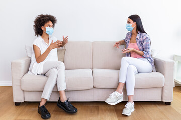 Two female best friends sitting in social distance wearing face mask and talking on the sofa, preventing covid 19 coronavirus pandemic infection spread.