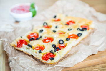 Focaccia with black olives, cherry tomatoes, rosemary and olive oil on a rustic wooden table. 