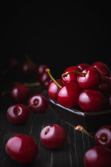 Vertical photo of ripe juicy cherries in a bowl on a rustic table with berries scattered around on a dark moody background with copy space for text.