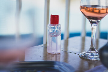 Bottle of hand sanitizer gel next to glass of rose wine, on restaurant table. People can wash their hands before eating.