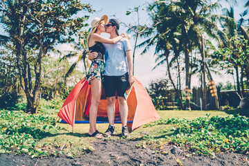 Young hipsters in romantic relationships spending weekends on camping enjoying recreation together, couple in love standing near tent kissing having active summer vacation trip in tropical environment