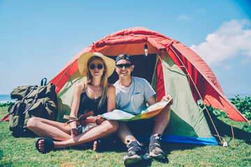 Portrait of hipster couple having adventure during journey camping on green grass recreating together,cheerful male traveler looking at camera while reading map and planning trip with girlfriend