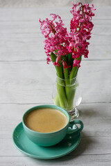 Pink hyacinth flowers in small transparent vase with coffee in blue coffee cup sitting on wooden surface. Hygge and still life concept.