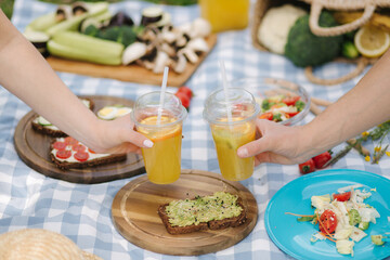 Photo of two hand hold orange lemonade in front of vegan picnic outdoors. Healthy food concept