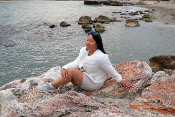Asian woman sits on the wild seashore and looks into the distance