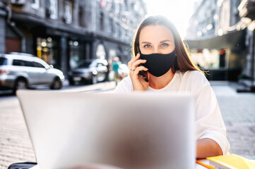 Obraz premium Close-up portrait of young woman with a protective mask at the table on the face on the summer terrace. Young woman works remotely with laptop in cafe, she looks at camera and talks on the smartphone
