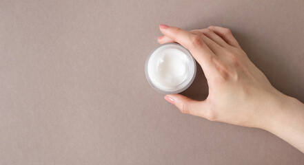Female hand holding cream moisturizer in glass jar on beige background and copy space. Beauty skincare concept