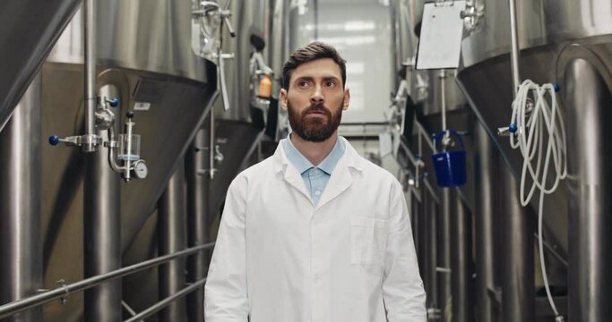Crop view of attentive young male worker inspecting equipment. Man in white lab coat walking through rows of steel brewing vats and looking aside.Concept of brewery and beer plant