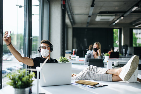 Young Man With Face Mask Back At Work Or School In Office After Lockdown, Taking Selfie.