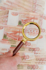 A man's hand holds a magnifying glass with a gold handle over a large number of Russian banknotes worth five thousand rubles