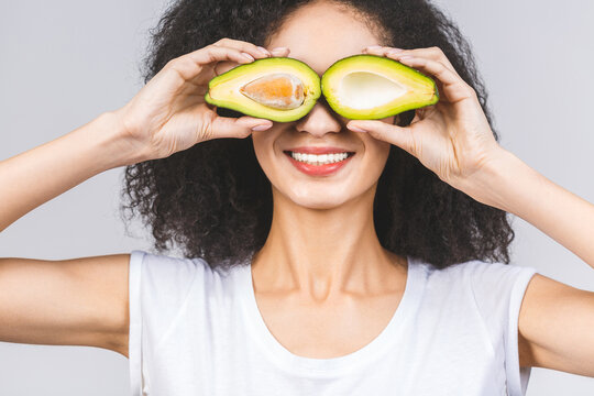 Pretty young woman holding an avocado in front of her face. Photo of smiling african american woman isolated on white or grey background. Beauty & Skin care concept.