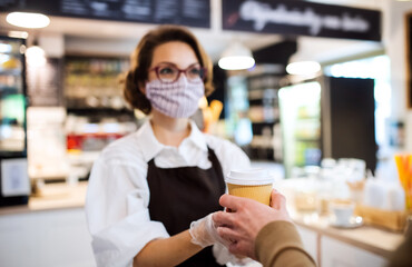 Young woman with face mask working indoors in coffee shop, serving customer.