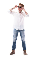 Young stylish handsome man in jeans and white shirt holding sunglasses looking at sunlight. Full length portrait isolated on white background. 