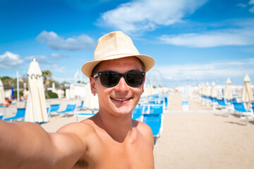 smiling young man with hat and sunglasses taking selfie with smartphone on the beach - smiling teen with martphone taking selfie