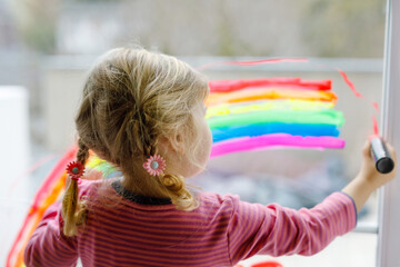 Adoralbe little toddler girl with rainbow painted with colorful window color during pandemic...
