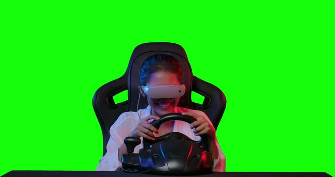Excited young asian teen gamer wearing vr glasses sitting in the racing seat simulator cockpit with steering wheel and playing in car racing game over green screen background. POV monitor screen view.