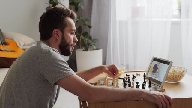 Modern Caucasian man playing chess and eating snacks with his online friend using tablet computer, self quarantine concept