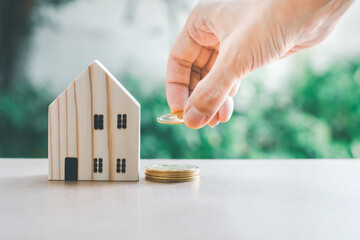 Saving money concept. Hand hold gloden coin with wooden model home on table with green bokeh background. Saving or investment for home in the future.