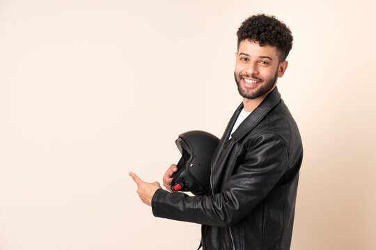 Young Moroccan man with a motorcycle helmet isolated on beige background pointing back
