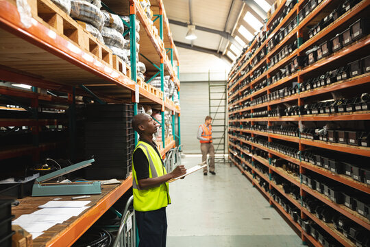 Black African American Male factory worker wearing a Hi-vis vest holding clipboard and checking stock at the factory