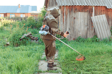 Asian man in a mosquito suit with a professional brush cutter trimmer mows the grass in the yard....