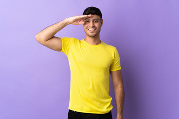 Young Colombian man isolated on purple background saluting with hand with happy expression