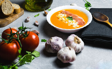 Salmorejo dish with eggs and ham, accompanied by tomato, oil, garlic and bread, along with a black cloth and a wooden spoon on a gray stone.
