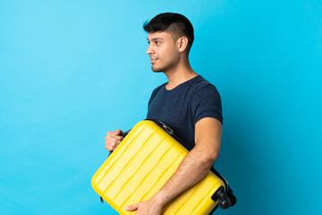 Young Colombian man isolated on blue background in vacation with travel suitcase