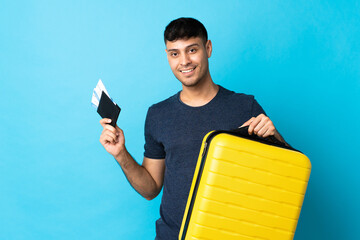Young Colombian man isolated on blue background in vacation with suitcase and passport