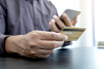 The businessman's hand is holding a credit card and using a smartphone for online shopping and internet payment in the office